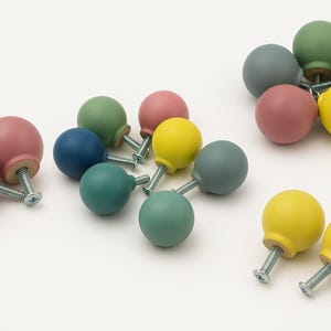 Könnte beinhalten: Eine Sammlung bunter, runder Schrankknöpfe in verschiedenen Farbtönen wie Rosa, Blau, Grün, Gelb und Grau. Jeder Knopf hat eine Metallschraube zur einfachen Montage. Die Knöpfe sind auf weißem Hintergrund angeordnet.