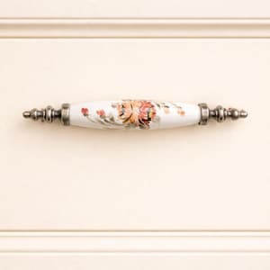 May include: A white ceramic cabinet pull with a floral design in shades of orange, pink, and green. The ends of the pull are a dark silver color. The pull is mounted on a cream-colored cabinet door.