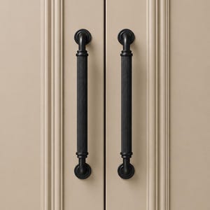 May include: Two vertical matte black cabinet pulls with a fluted, ribbed texture. Each handle features a cylindrical center bar with vertical grooves and rounded mounting bases at the top and bottom, attached to light beige paneled cabinet doors.