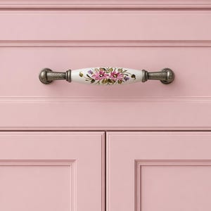 May include: A pink cabinet with a decorative handle. The handle is white with a floral design in pink and purple, and the hardware is a dark, antique silver colour. The cabinet doors are a solid, light pink. The handle is approximately 15 cm long.