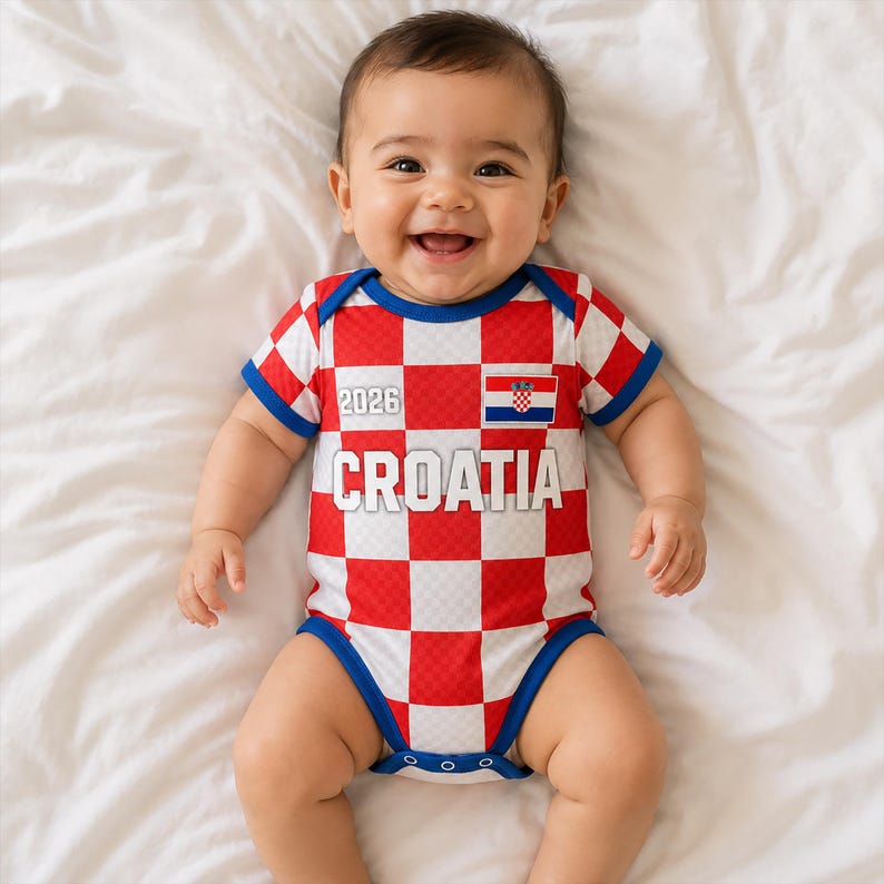 K&ouml;nnte beinhalten: Ein kurz&auml;rmeliger Baby-Body mit einem rot-wei&szlig;en Karomuster. Das Kleidungsst&uuml;ck zeigt den Schriftzug 'CROATIA' in gro&szlig;en wei&szlig;en Buchstaben, die Zahl '2026' in kleinerer wei&szlig;er Schrift und ein kleines rechteckiges kroatisches Flaggensymbol auf der Brust.