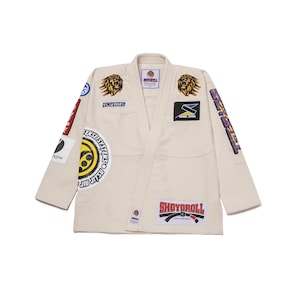 May include: Cream-colored Jiu-Jitsu gi with multiple patches. Patches include lion graphics, the word "COMPETITOR", and the text "SHOYOROLL". The gi has a V-neck design and is displayed on a white background.