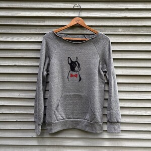 May include: A heather gray long-sleeve sweatshirt featuring a graphic of a black and white Boston Terrier dog wearing a red bow tie with white polka dots. The garment has a wide neckline, long sleeves with ribbed cuffs, and a front kangaroo pocket.