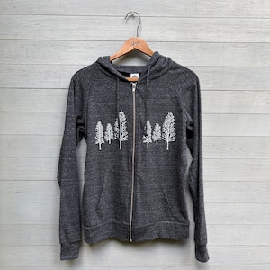May include: A charcoal gray zip-up hoodie featuring a white graphic print of a forest of pine trees across the chest. The garment includes a hood with matching drawstrings, long sleeves, and two front pockets, hanging on a wooden hanger.