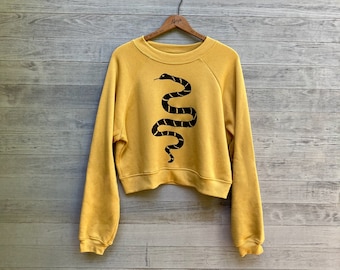 May include: A mustard yellow sweatshirt with a black snake graphic on the front. The sweatshirt has a relaxed fit and long sleeves.