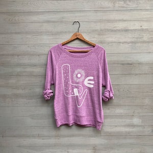 May include: A purple sweatshirt with a white graphic of the word "LOVE" with a stylized design. The letters are filled with small white dots.