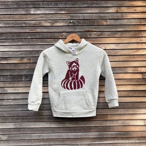 May include: A light gray hoodie with a maroon red panda graphic. The hoodie has a kangaroo pocket and a hood. The red panda is sitting with its tail curled up. The hoodie is hanging on a wooden hanger.