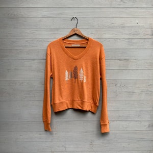 Könnte beinhalten: Orangefarbener Pullover mit langen Ärmeln, V-Ausschnitt und einem Waldmotiv auf der Vorderseite.
