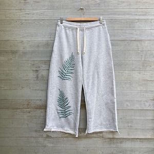 Könnte beinhalten: Graue Jogginghose mit Kordelzug und zwei grünen Farnblatt-Designs. Die Hose hat einen rohen Saum und hängt an einem Holzbügel vor einem grauen Holzgrund.