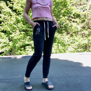 Könnte beinhalten: Eine Frau trägt eine schwarze Jogginghose mit einem gestickten Eulenmotiv auf der linken Tasche und ein pinkfarbenes, bauchfreies T-Shirt mit einem Wassermelonen-Motiv. Sie trägt schwarze Schlappen.