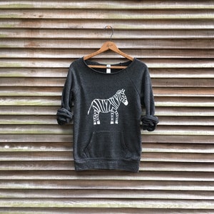 May include: A dark gray sweatshirt with a white zebra graphic print. The sweatshirt has a crew neck and a kangaroo pocket.