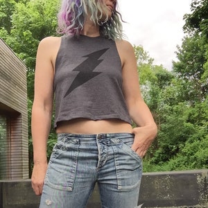May include: A woman wearing a gray cropped tank top with a black lightning bolt graphic and blue jeans with distressed details.