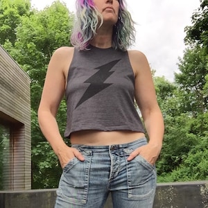 May include: A woman wearing a gray cropped tank top with a black lightning bolt graphic and blue denim jeans with distressed details.