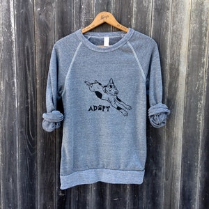 May include: A heather gray sweatshirt with a black graphic of a dog leaping and the word "ADOPT" printed below. The long sleeves are rolled up. The sweatshirt is hanging on a wooden hanger against a weathered wood background.