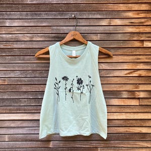 May include: A light green tank top with a floral design. The sleeveless top features a crew neck and a row of black line-drawn flowers across the front. The tank top is hanging on a wooden hanger against a wooden background.
