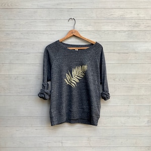 Puede incluir: Una sudadera gris oscuro con un diseño de helecho dorado en la parte delantera. La sudadera está colgada de una percha de madera.