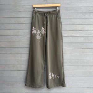 K&ouml;nnte beinhalten: Olivgr&uuml;ne weit geschnittene Jogginghose mit elastischem Bund und Kordelzug. Die Vorderseite der Hose ist mit wei&szlig;en Strichzeichnungen verschiedener Farne und botanischer Bl&auml;tter auf der oberen rechten und unteren linken Seite verziert.