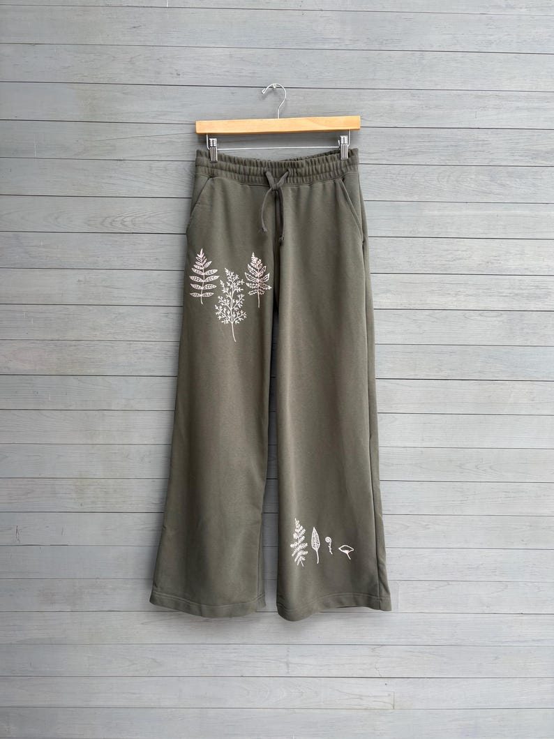 K&ouml;nnte beinhalten: Olivgr&uuml;ne weit geschnittene Jogginghose mit elastischem Bund und Kordelzug. Die Vorderseite der Hose ist mit wei&szlig;en Strichzeichnungen verschiedener Farne und botanischer Bl&auml;tter auf der oberen rechten und unteren linken Seite verziert.