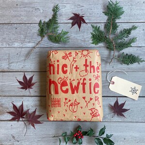 May include: A brown paper gift bag with red illustrations of a cat, a fox, a frog, a fence, and the words "nic + the newfie". The bag is surrounded by green and red foliage, including holly berries.