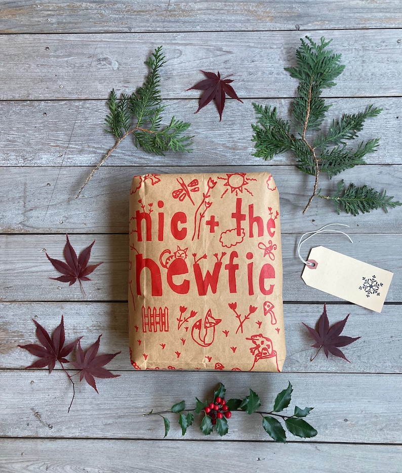 May include: A brown paper gift bag with red illustrations of a cat, a fox, a frog, a fence, and the words "nic + the newfie". The bag is surrounded by green and red foliage, including holly berries.