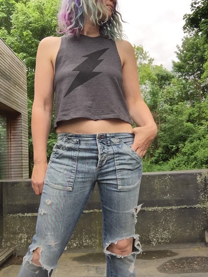 May include: A woman wearing a gray cropped tank top with a black lightning bolt graphic and blue jeans with distressed details.