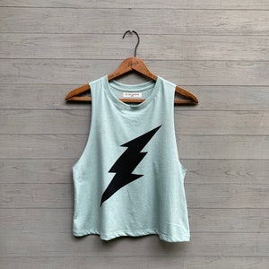 May include: A light blue sleeveless tank top with a black lightning bolt graphic. The tank top is hanging on a wooden hanger.
