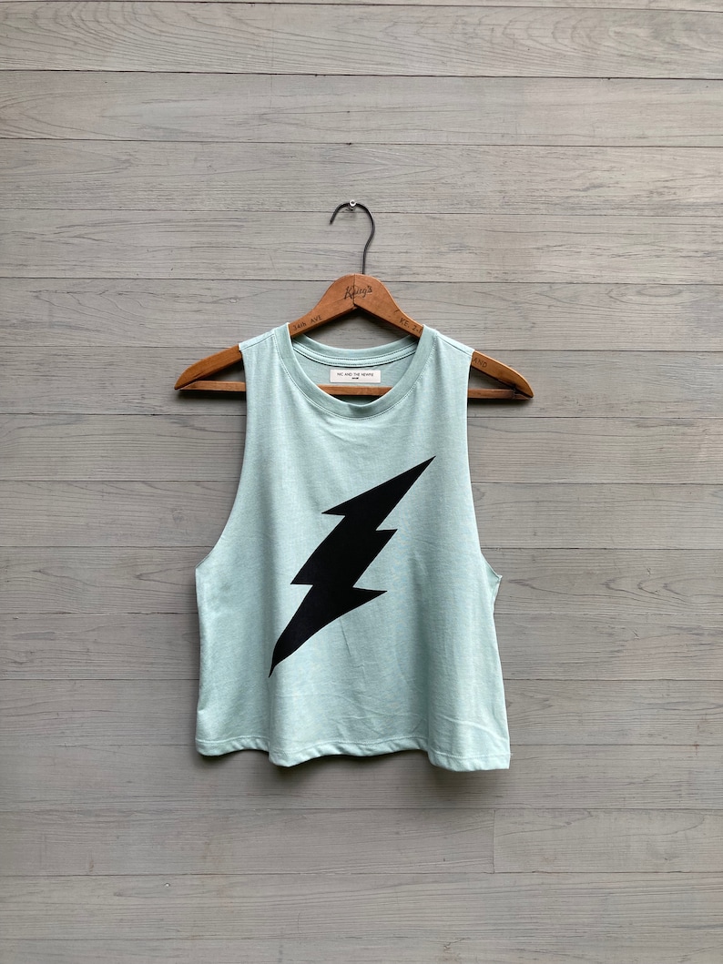 May include: A light blue sleeveless tank top with a black lightning bolt graphic. The tank top is hanging on a wooden hanger.