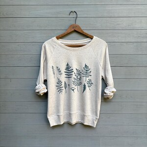 Puede incluir: Una sudadera color crema con cuello redondo y mangas largas, con un estampado botánico verde oscuro de varias hojas y plantas en el pecho. Las mangas están enrolladas. La sudadera cuelga de una percha de madera sobre un fondo de madera gris.