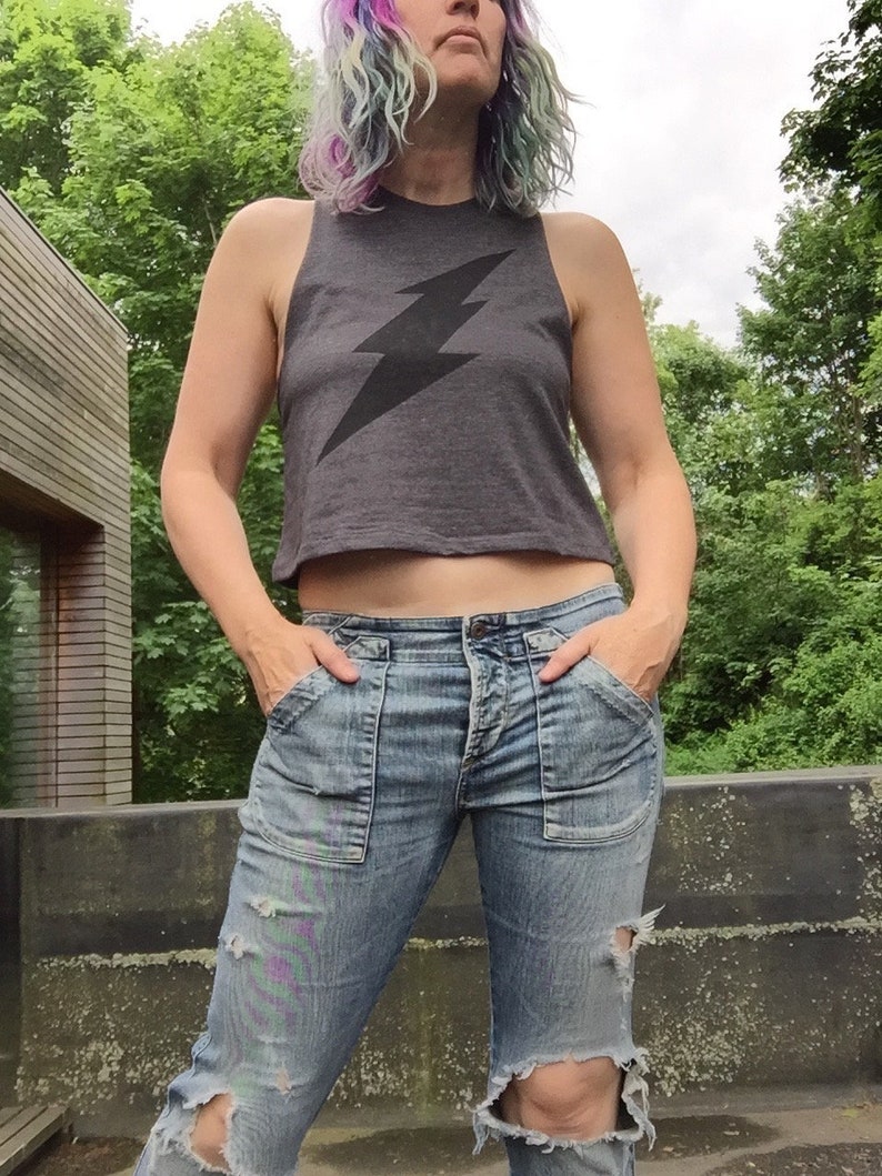 May include: A woman wearing a gray cropped tank top with a black lightning bolt graphic and blue denim jeans with distressed details.