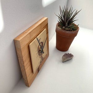 May include: A wooden clock with a skeleton illustration on the face. The clock is on a white surface with a small potted plant and a purple stone.