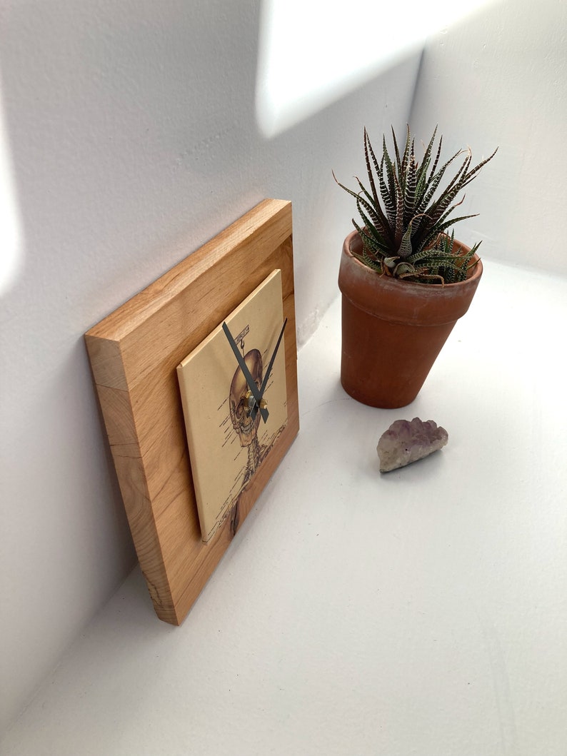 May include: A wooden clock with a skeleton illustration on the face. The clock is on a white surface with a small potted plant and a purple stone.