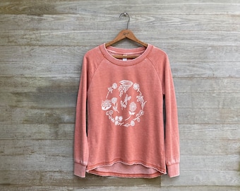 May include: A long-sleeved, crew-neck sweatshirt in a faded, washed-out terracotta orange color. The front features a circular graphic composed of various white line-art floral and botanical illustrations. The hem has a raw, unfinished edge.