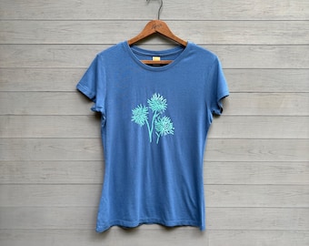May include: A blue short-sleeved t-shirt with a floral design in a lighter shade of blue. The shirt is hanging on a wooden hanger against a gray wooden background. The floral design is centered on the front of the shirt.
