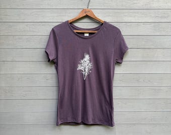 May include: A short-sleeved, purple t-shirt with a white tree graphic on the front. The t-shirt is hanging on a wooden hanger against a gray wooden background. The tree graphic is a detailed, stylized illustration.