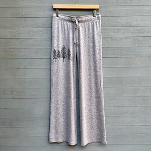 May include: Light gray wide-leg sweatpants with a drawstring waist, hanging on a wooden hanger. A black graphic of stylized trees is printed on the left leg. The pants are made of a soft, comfortable material.