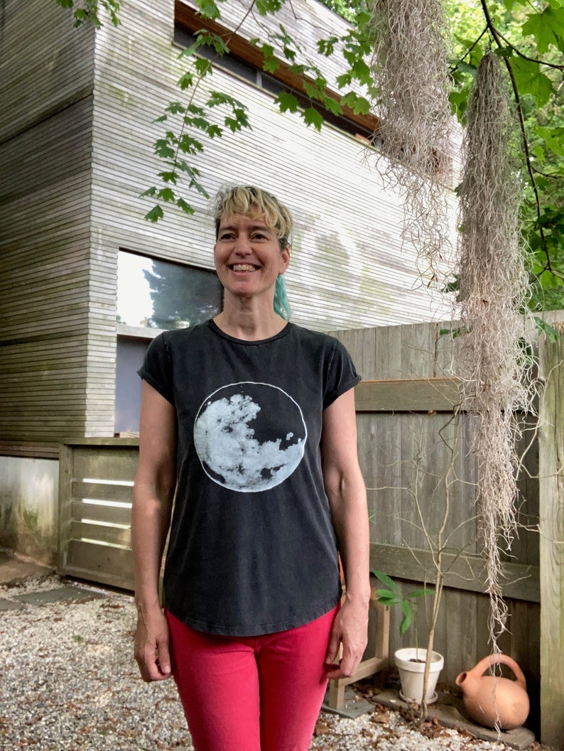 K&ouml;nnte beinhalten: Eine Frau mit kurzen, blauen Haaren tr&auml;gt ein schwarzes T-Shirt mit einem wei&szlig;en Mond-Motiv. Sie tr&auml;gt eine rote Hose und steht vor einem Holzzaun.