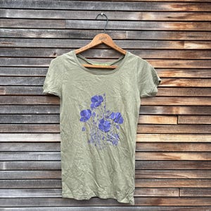 Pu&ograve; includere: T-shirt verde oliva a maniche corte con scollo tondo. La maglietta presenta un motivo floreale di fiori viola. La maglietta &egrave; appesa a una gruccia di legno su uno sfondo di assi di legno.