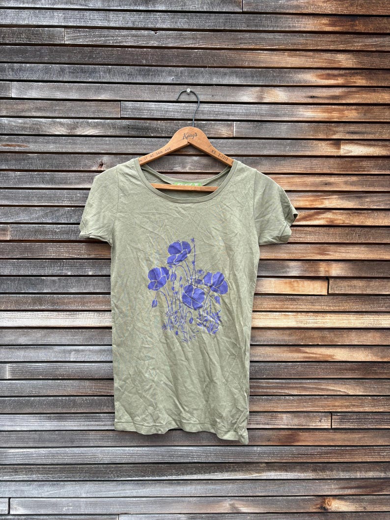 Pu&ograve; includere: T-shirt verde oliva a maniche corte con scollo tondo. La maglietta presenta un motivo floreale di fiori viola. La maglietta &egrave; appesa a una gruccia di legno su uno sfondo di assi di legno.