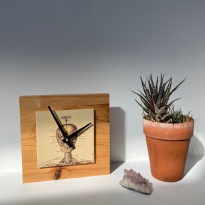 May include: A wooden clock with a skeleton head design on the face. The clock is sitting on a wooden base and is next to a small potted succulent plant and a purple crystal.