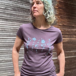 Op de afbeelding: Een paars T-shirt met korte mouwen en een lichtblauw bloemmotief. Het shirt wordt gecombineerd met een rode broek. De achtergrond is een houten muur.