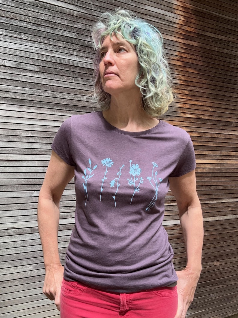 Op de afbeelding: Een paars T-shirt met korte mouwen en een lichtblauw bloemmotief. Het shirt wordt gecombineerd met een rode broek. De achtergrond is een houten muur.