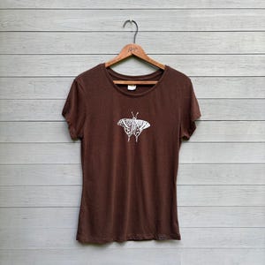 May include: A short-sleeved brown t-shirt featuring a white graphic print of a butterfly centered on the chest. The shirt has a round neckline and is displayed on a wooden hanger, showcasing a simple design with a solid dark brown fabric base.