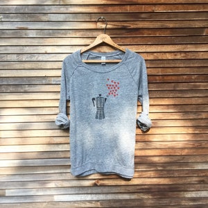 May include: A heather gray long-sleeve sweatshirt featuring a black coffee maker graphic with red heart accents. The sweatshirt is displayed on a wooden hanger against a wooden plank background. The sleeves are rolled up.