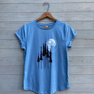 Puede incluir: Una camiseta azul claro con un gr&aacute;fico de una luna llena y cinco &aacute;rboles negros.