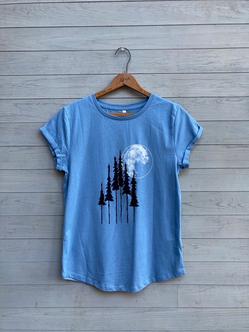 Puede incluir: Una camiseta azul claro con un gr&aacute;fico de una luna llena y cinco &aacute;rboles negros.