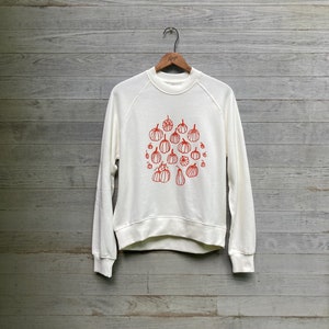 Op de afbeelding: Een wit sweatshirt met een rood pompoenontwerp. De pompoenen zijn van verschillende groottes en vormen en zijn in een cluster op de voorkant van het sweatshirt gerangschikt.