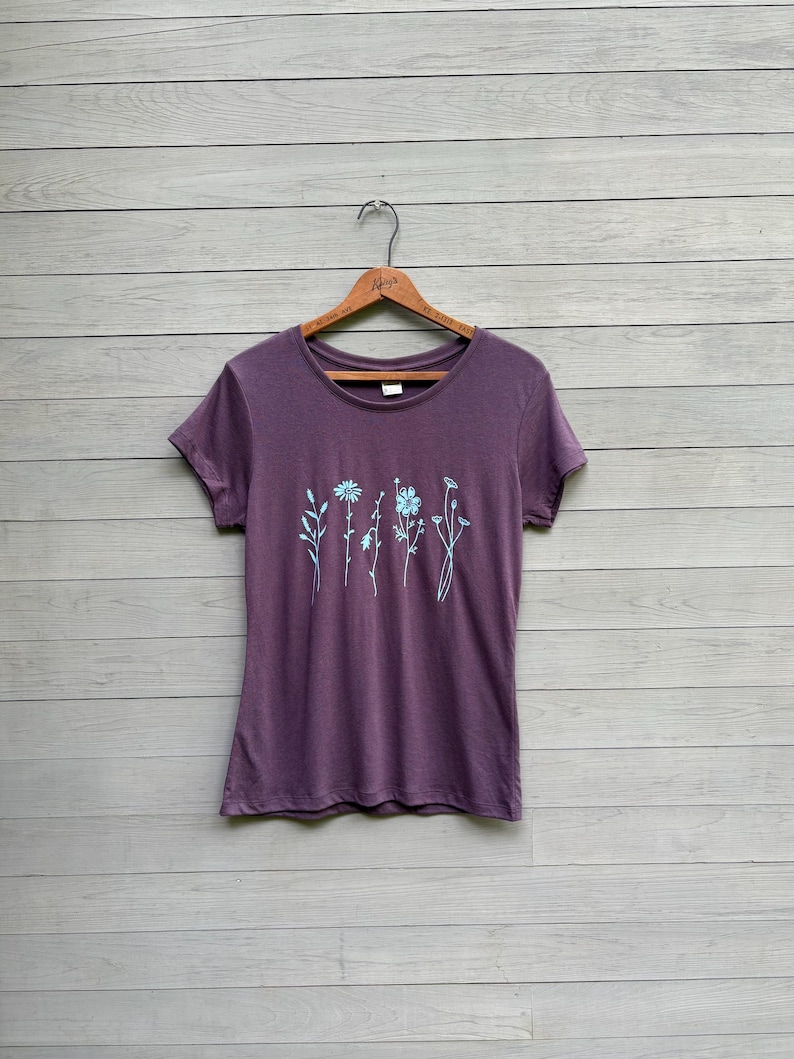 Op de afbeelding: Een paars T-shirt met een ronde hals en korte mouwen. Het shirt heeft een lichtblauw bloemenontwerp op de voorkant. Het hangt aan een houten hanger tegen een grijze houten achtergrond.