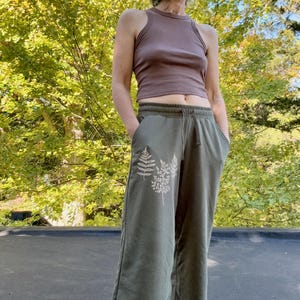 K&ouml;nnte beinhalten: Ein &auml;rmelloses, mauvefarbenes bauchfreies Oberteil mit Stehkragen und eine olivgr&uuml;ne Jogginghose mit wei&szlig;em Farnblatt-Aufdruck auf der Vorderseite. Die Hose hat einen elastischen Bund mit Kordelzug sowie seitliche Eingrifftaschen.