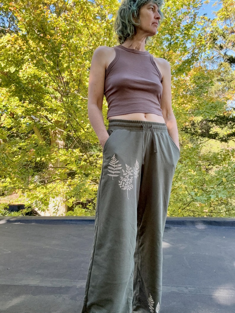 K&ouml;nnte beinhalten: Ein &auml;rmelloses, mauvefarbenes bauchfreies Oberteil mit Stehkragen und eine olivgr&uuml;ne Jogginghose mit wei&szlig;em Farnblatt-Aufdruck auf der Vorderseite. Die Hose hat einen elastischen Bund mit Kordelzug sowie seitliche Eingrifftaschen.