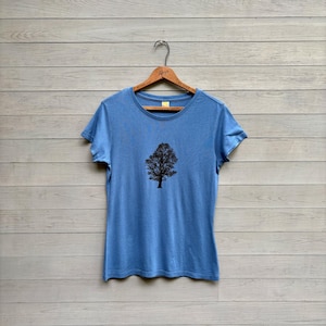 May include: A light blue t-shirt with a crew neck and short sleeves. The shirt features a black graphic of a tree centered on the front. The t-shirt is hanging on a wooden hanger against a light gray wooden background.